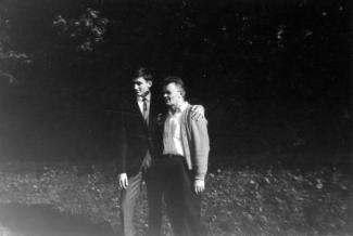 Photo en noir et blanc d’un jeune homme tenant son père par les épaules. Ils sont à l’extérieur, des arbres derrière eux. 