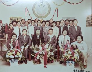 hoto couleur un peu délavée montrant un groupe de 24 personnes d’origine sino-cambodgienne réparties sur deux rangées prenant la pose de façon officielle, avec trois grands paniers de fleurs devant eux.