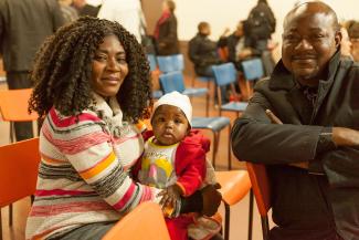 Une femme assise tient un enfant dans ses bras. Elle fait face à la caméra, tout comme un homme assis à côté d’elle.