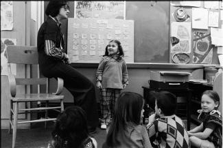 Une élève est debout dos au tableau, à côté de son enseignante. En arrière-plan, quatre élèves.