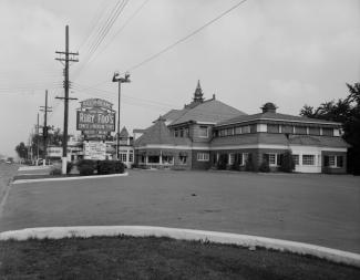 Photographie en noir et blanc d’un bâtiment le jour. À gauche, l’enseigne mentionne le nom du restaurant. 