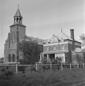 Photo en noir et blanc d’une église et de son presbytère dans un environnement agricole.