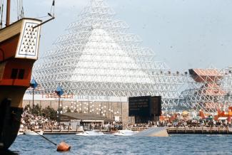 Vue extérieure du lac des Dauphins et de l’attraction nommée Gyrotron de l’Expo 67, avec sa pyramide blanche et le volcan à droite de la photographie. Le Minirail rejoignant les deux structures est également visible. 