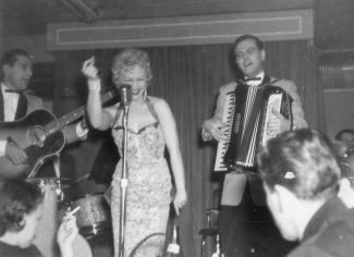 Photo en noir et blanc d’une chanteuse et de ses musiciens, un accordéoniste et un guitariste, sur la scène d’un cabaret. 