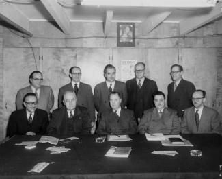 Photographie en noir et blanc d’un groupe d’hommes se tenant derrière une table de travail. Ils portent tous un complet-cravate.