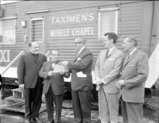 Photographie en noir et blanc montrant cinq hommes debout devant une roulotte sur laquelle on peut lire “Taximen’s mobile chapel”.