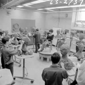 Photo en noir et blanc montrant un cours de sculpture dans un grand atelier éclairé. Une femme est le modèle, assise au centre sur un petit podium. Des élèves travaillent debout chacun devant une petite table tout autour d’elle.