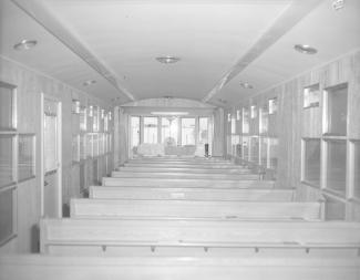 Photo en noir et blanc montrant l’intérieur d’une grande roulotte transformée en chapelle avec une série de bancs en bois et, au fond, un autel.
