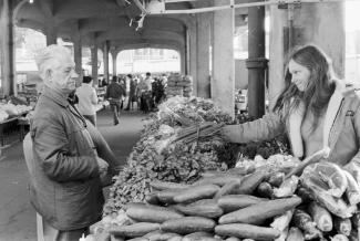 Photo en noir et blanc d’un homme âgé se tient devant un kiosque de légumes dans un marché. 