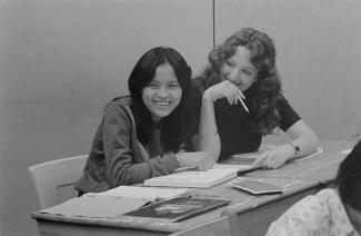 Deux femmes, assises à leurs pupitres souriant. Des livres sont ouverts sur les pupitres.