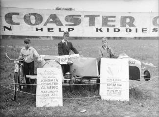 Trois garçons prennent la pose à côté de leurs boîtes à savon et deux affiches en anglais devant eux annoncent une course. 
