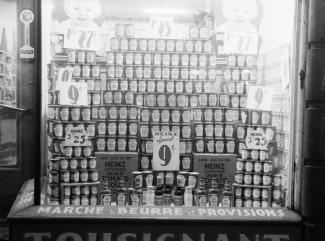 Photographie noir et blanc d’une vitrine avec un étalage de produits en conserve de la marque Heinz. Sur le rebord de la vitrine, il est écrit « Marché à beurre et provisions ».