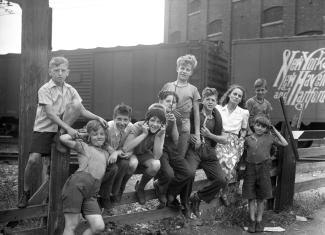 La femme de lettres Gabrielle Roy entourée de neuf petits garçons du quartier Saint-Henri à Montréal. Ils sont assis sur une clôture au coin des rues Saint-Augustin et Saint-Ambroise près de la voie ferrée.