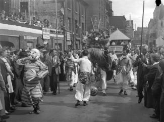 La communauté chinoise célèbre le jour de la Victoire par un défilé dans les rues du Quartier chinois de Montréal. La scène présente la procession du lion et un homme vêtu d’un masque de théâtre chinois. 