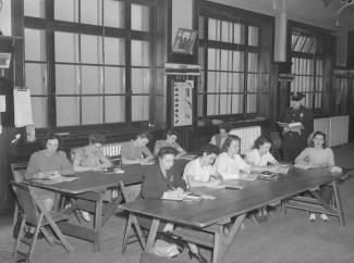 Photographie en noir et blanc de dix femmes policières assises dans une salle de classe avec un policier debout tenant un livre.  