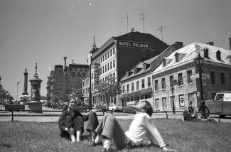 Photo en noir et blanc de la place Jacques-Cartier, prise juste au sud de la rue Saint-Paul vers la rue Notre-Dame. Deux jeunes sont assis à l’avant-plan dans l’herbe et un est couché.