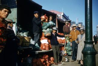 Photo couleur montrant des maraîchers et une femme prenant la pose devant leur étal remplis de sacs de légumes.