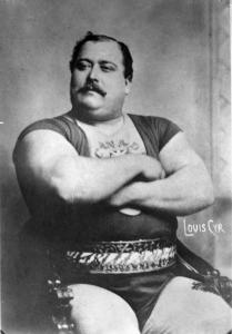 Photographie en noir et blanc d’un homme portant un costume de lutteur sur lequel il est écrit « Canada » au niveau du torse. L’homme a les bras croisés et porte une moustache. 