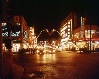 Photo couleur montrant la Plaza Saint-Hubert décorée pour le temps des fêtes en soirée avec les enseignes lumineuses, des voitures et des gens au coin de la rue sur le trottoir.