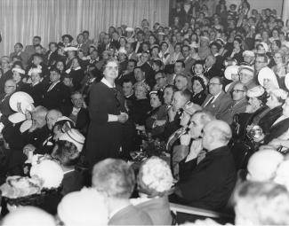 Une femme, les mains jointes, debout au milieu d’une assemblée de personnes assises, regarde vers sa droite.
