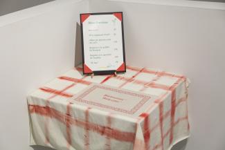 Photo d’une oeuvre artistique montrant une table, un napperon et un menu intitulé « Menu d’exotisme ».