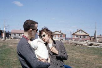 Un homme tenant un bébé dans ses bras et une femme qui regarde le bébé se tiennent debout sur un vaste terrain dans un nouveau quartier en construction. À l’arrière-plan, différents modèles de maisons unifamiliales.