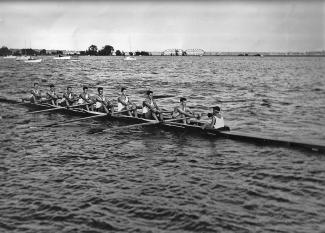 Photo en noir et blanc d’un bateau d’aviron à huit rameurs et un barreur leur faisant face avec un porte-voix. En arrière plan, les arches d’un pont ferroviaire. 