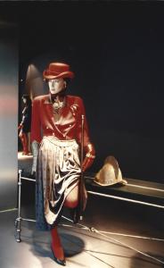 Vitrine de mode automnale avec un mannequin féminin à l’allure déterminée portant un gant d’armure métallique. Il porte un court veston rouge sur une longue jupe imprimée et s’appuie contre une console d’étain et de verre où repose un ancien casque milita