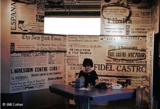 Une hôtesse assise devant une table avec derrière elle un mur tapissé d’articles de journaux évoquant différents moments de la révolution cubaine.