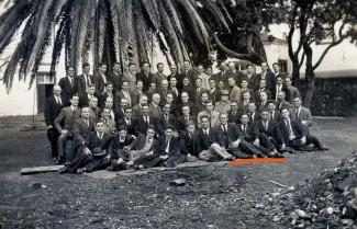 Fotografia a preto e branco que mostra um grupo de homens com colete e gravata repartidos em cinco filas, a fazer pose em frente a um edifício e uma palmeira.