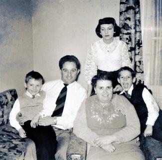 Manuel Jácome, uma criança sentada ao seu colo, uma mulher e uma criança à direita e uma mulher de pé atrás deles, numa sala de estar.
