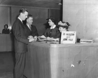 Deux hommes sont debout devant un comptoir, derrière lequel deux femmes sont debout. Les deux femmes portent un chapeau. Écriteau sur le comptoir : « Protection de la jeune fille ». Au loin, une femme à un comptoir parle au téléphone. 