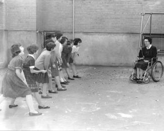 Des fillettes sont dehors et rient, elles sont en position de départ pour courir. Devant elle, une femme en fauteuil roulant.