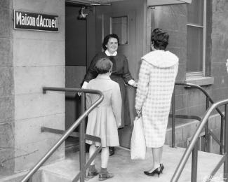 Une femme debout tient une porte ouverte pour une femme et un enfant que l’on voit de dos. Sur la gauche, une plaque où est écrit : « Maison d’Accueil ».