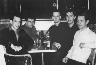 Cinq jeunes hommes sont assis dans un bar autour d’une petite table remplie de bouteilles et de verres.