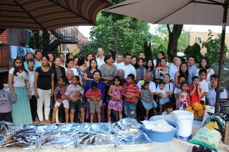 Photo d’une famille d’une cinquantaine de personnes de différentes générations dans un cour avec des plats de nourriture recouverts en avant-plan. 