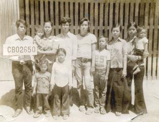 Photo en noir et blanc d’un groupe d’adultes et d’enfants cambodgiens. Un adulte tient une pancarte avec un numéro. 