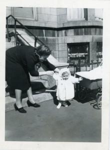 Photo en noir et blanc d’un bébé qui semble faire ses premiers pas sur le trottoir devant la maison avec sa mère qui le tient par la main. Le landau de l’enfant se trouve à droite.  