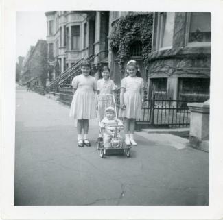 Trois filles d’une dizaine d’années accompagnées d’un bébé en poussette posent devant une rangée de maisons en pierres grises.