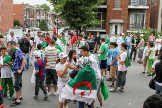 Une foule arborant le drapeau de l'Algérie. En arrière-plan, des maisons