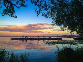 Scène au bord de l’eau avec un ciel de soirée aux couleurs chaudes et neuf personnes assises sur un quai.