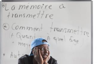 Un garçon portant une casquette se tient la tête entre les mains. Derrière lui, un tableau sur lequel on peut lire "La mémoire à transmettre"