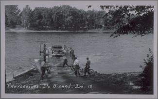 Carte postale en noir et blanc montrant un traversier accosté avec une voiture à son bord et trois cyclistes qui se préparent à embarquer. On voit un quatrième homme devant le traversier.