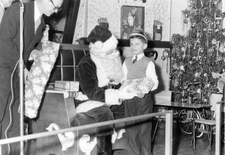 Photo en noir et blanc d’un enfant avec le père Noël qui lui remet un cadeau.