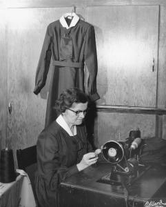 Une femme, concentrée sur son ouvrage, coud manuellement un vêtement. Elle est assise devant une machine à coudre. Derrière elle, un costume est suspendu, elle porte le même. 