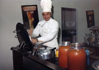 Un homme habillé en blanc avec chapeau de chef brasse un immense chaudron. Sur le comptoir, deux gros pots de sauce tomates. 
