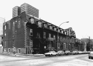 Photo en noir et blanc d’une rangée de triplex avec des voitures stationnées devant, prise du coin de la rue.