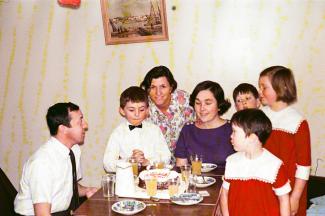 Trois adultes et quatre enfants sont autour d’une table où se trouve un gâteau d’anniversaire et des jus.