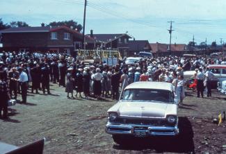 Photo couleur d’un événement réunissant une foule nombreuse dans un nouveau quartier résidentiel. Une voiture se trouve à l’avant-plan.