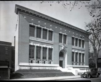 Photographie en noir et blanc montrant un bâtiment imposant de forme rectangulaire avec une arche autour de la porte et un toit entouré d’une corniche ornementée. Une voiture noire est stationnée devant le bâtiment.